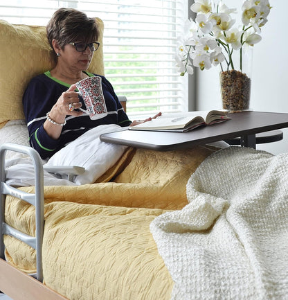 Lady lying in bed using Invacare's item number 6417, Hospital-Style, Brown Overbed Table reading a book and holding a mug.